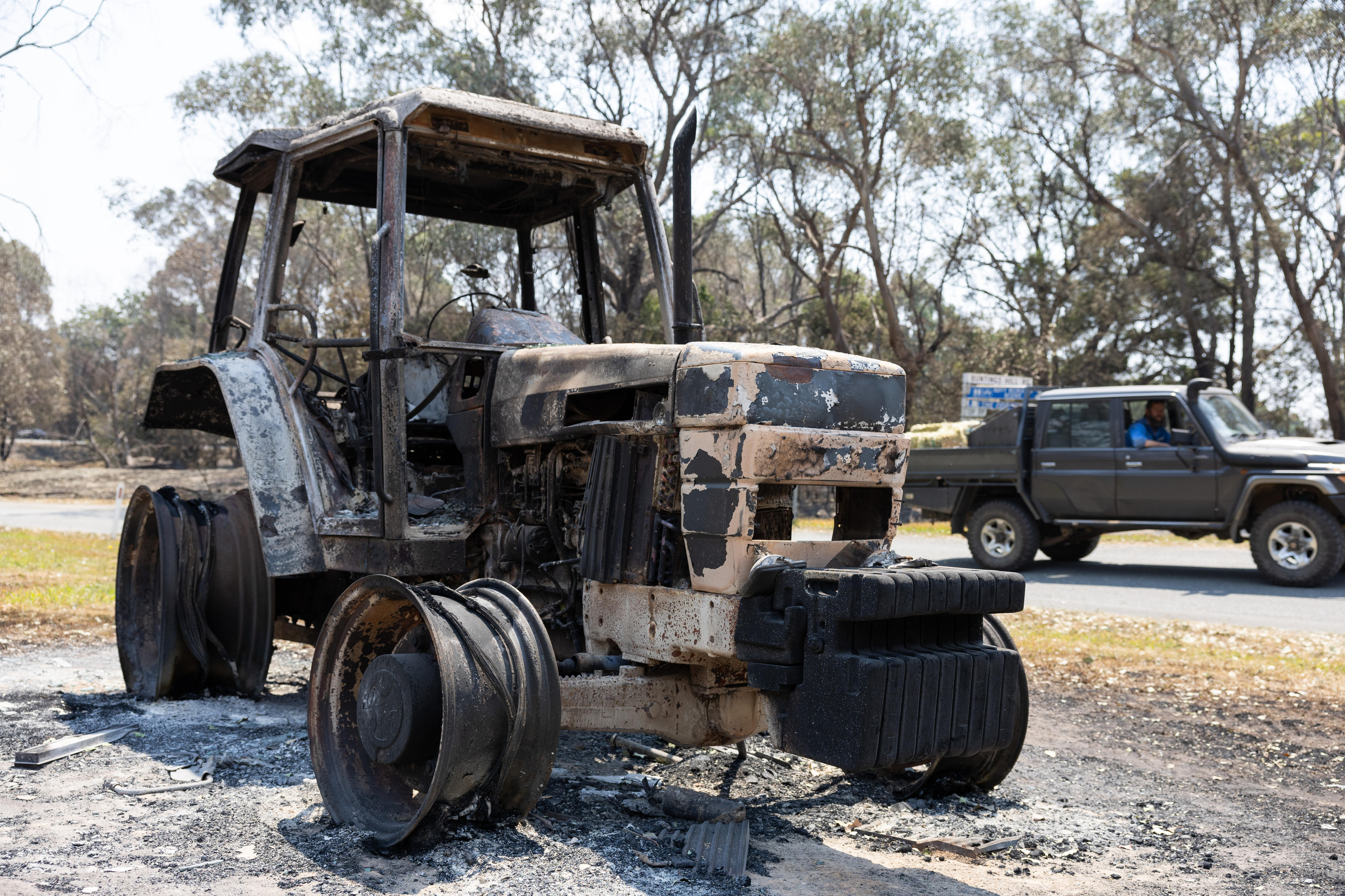 Un tractor quemado con un camión al fondo y el conductor mirándolo.