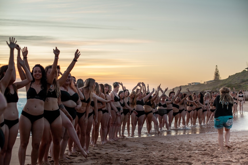 A huge line of laughing women in their underwear pose for a group photoshoot on the beach at dusk