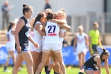 A group of AFLW teammates huddle together after a win, while their opponents look dejected.