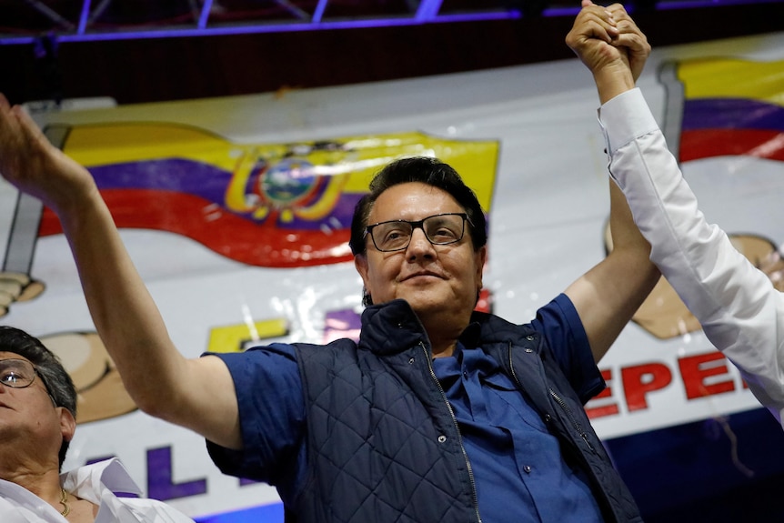 A man in a blue vest and blue shirt holds his hands in the air, at a rally