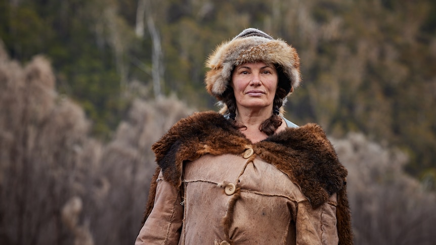 A woman in her 50s in a possum skin cloak and hat, two brown plaits, standing in front of forested hills