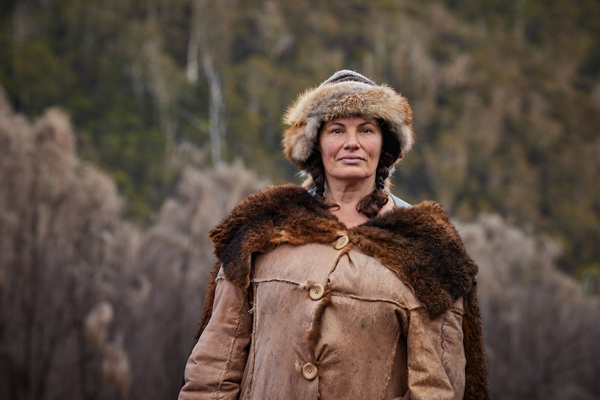 A woman in her 50s in a possum skin cloak and hat, two brown plaits, standing in front of forested hills