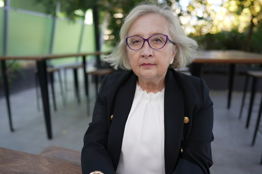 A middle-aged white woman with a black blazer and glasses sitting in a courtyard 