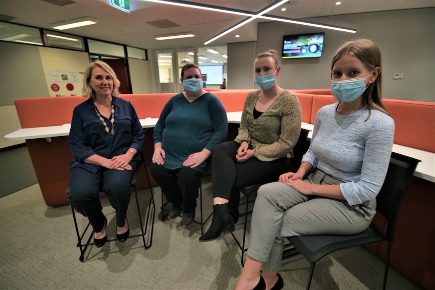 Four women sit in a university space, three are wearing face masks