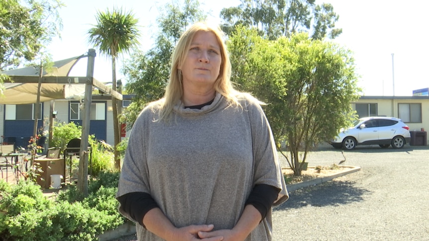 A woman in a beige shawl stands in front of a garden in a carpark. She looks worried.