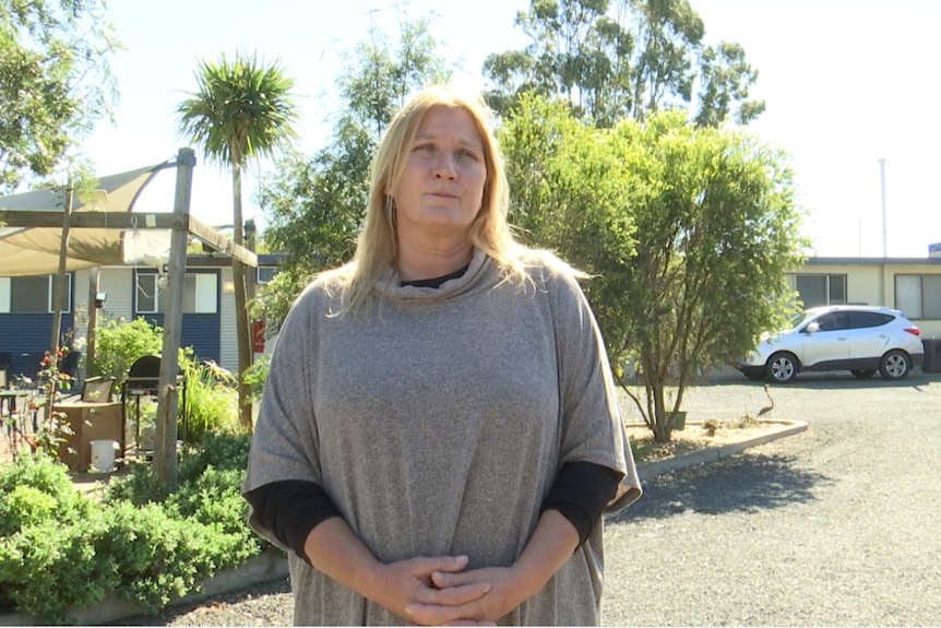 A woman in a beige shawl stands in front of a garden in a carpark. She looks worried.