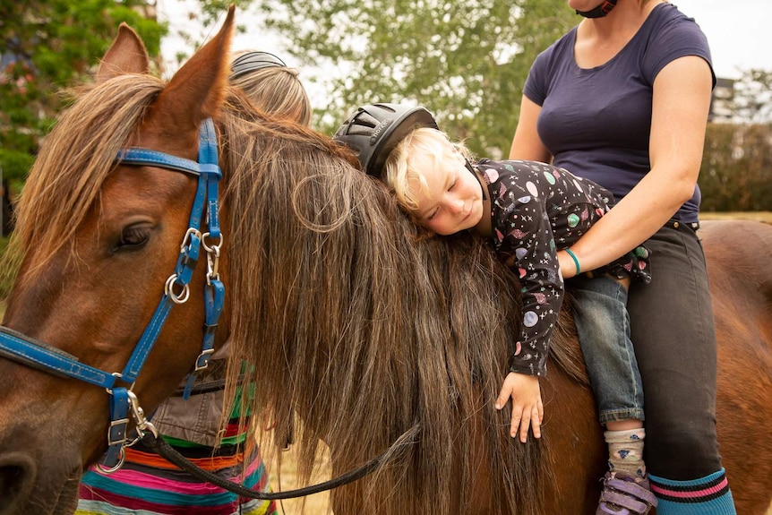 A young child is saddled up on a brumby with her mother.