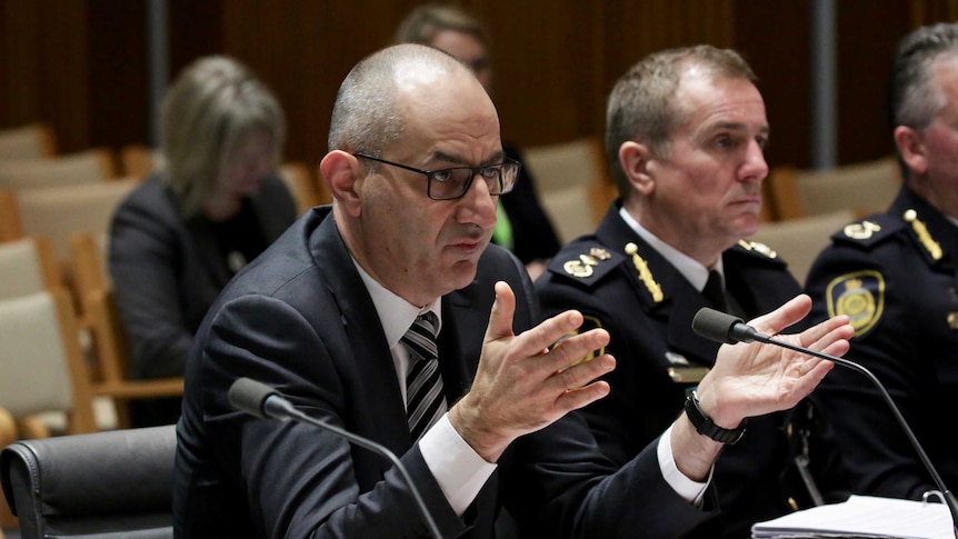 Mike Pezzullo holds his hands in the air as he sits at a desk inside a Parliament House committee room