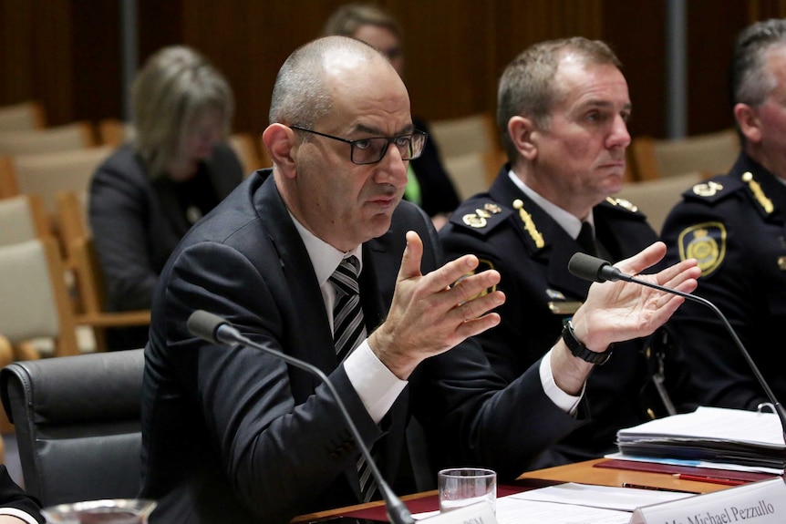 Mike Pezzullo holds his hands in the air as he sits at a desk inside a Parliament House committee room