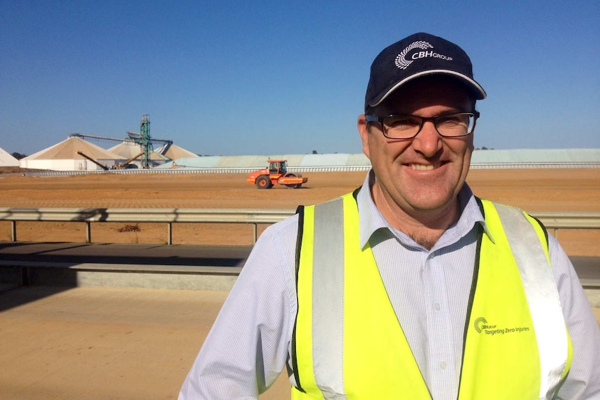 Man with a cap standing in front of the site where crops are received, with a tractor and silos in background.