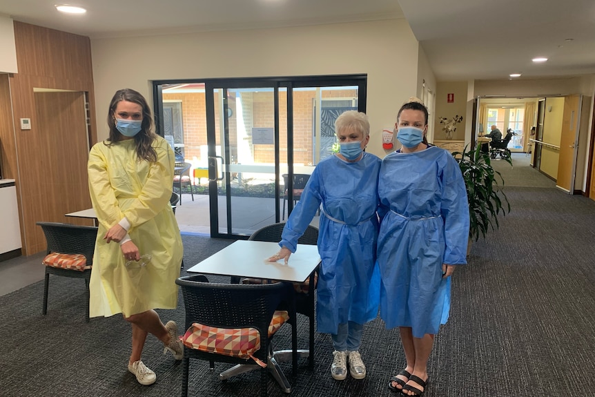 Three women dressed in PPE clothing stand separated by a table