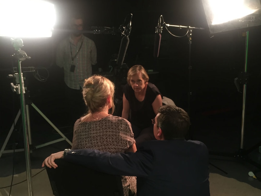 Journalist leaning forward to a woman sitting on a chair in a TV studio with a man next her putting his arm around her.