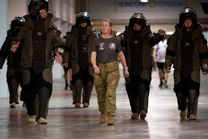 A woman wearing a t-shirt and camouflague pants walks with a group of people wearing black bomb disposal suits.