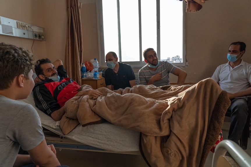 A man reclines in a hospital bed, smiling and talking to men and boys visiting him