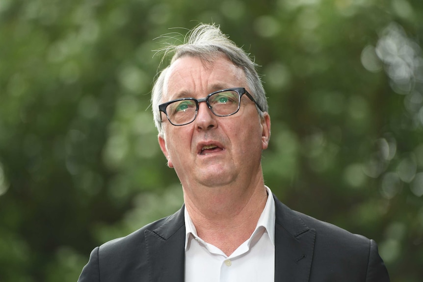 A grey-haired man in a suit with no tie speaking at a media conference