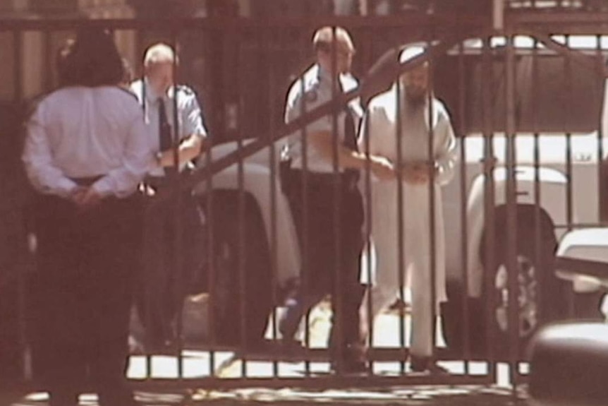 Man in white robe with white cap being led by police officers past gates.