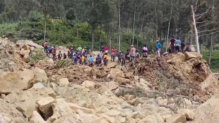 People are seen at a distance standing on rocks and rumble from a landslide. 