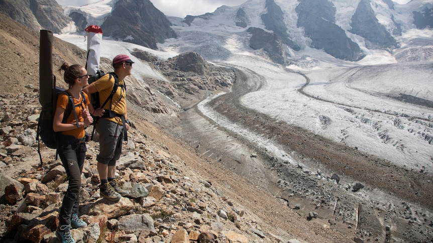 Two people with backpacks on stand on rocks and look down at the snow on the ground and alps in the background. 