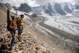 Two people with backpacks on stand on rocks and look down at the snow on the ground and alps in the background. 