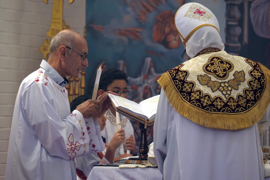 A Coptic Orthodox priest, and man, both in white church robes, conducting a ceremony during a church service.