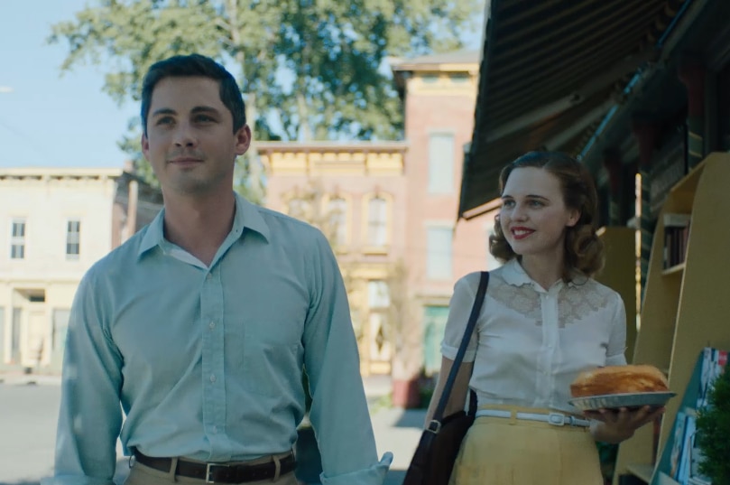 A man with short dark hair in blue button up shirt walks down street with woman in white blouse and yellow skirt holding cake.