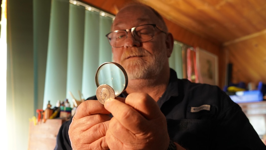 A man holds a magnifying glass up to a small silver medal