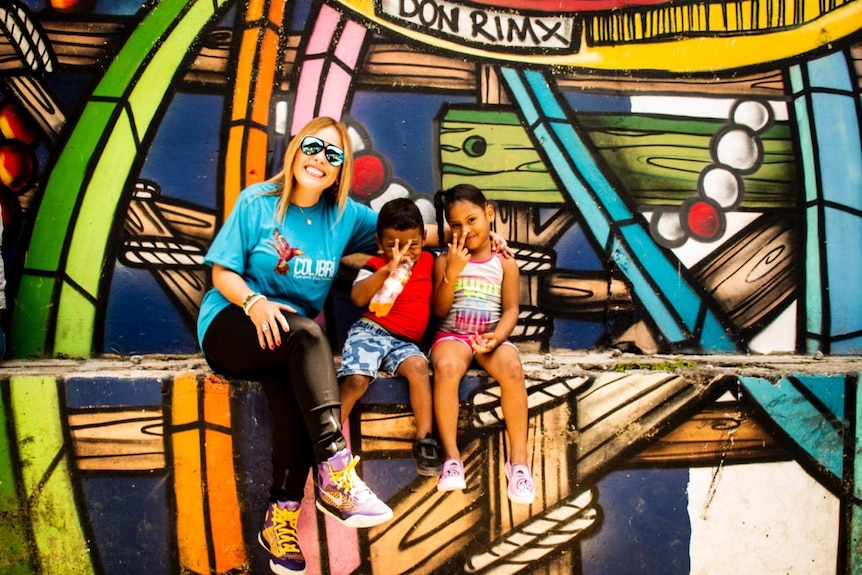 A woman with blonde hair and reflective glasses smiles next to two young kids. A colourful mural can be seen in the background.