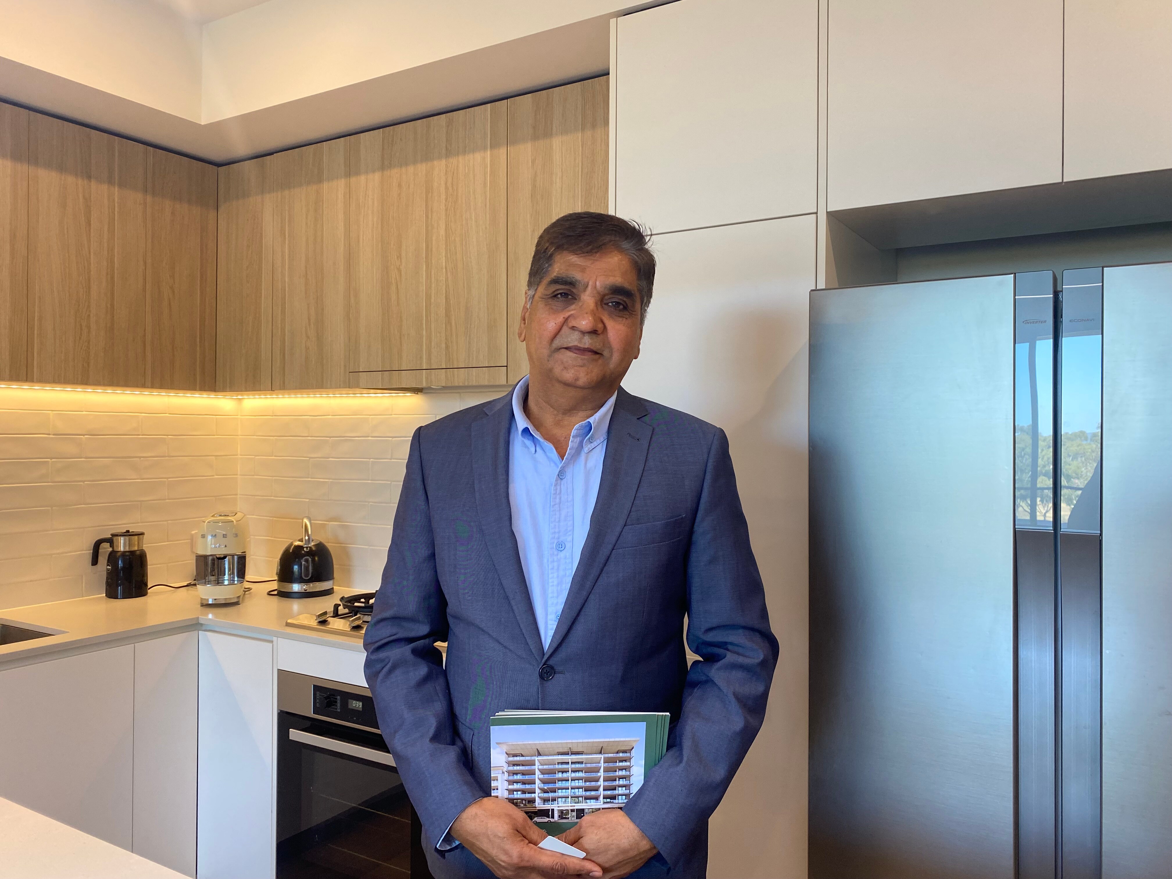 An older Indian man wearing a suit and collared shirt without a tie stands in the kitchen of a modern house.