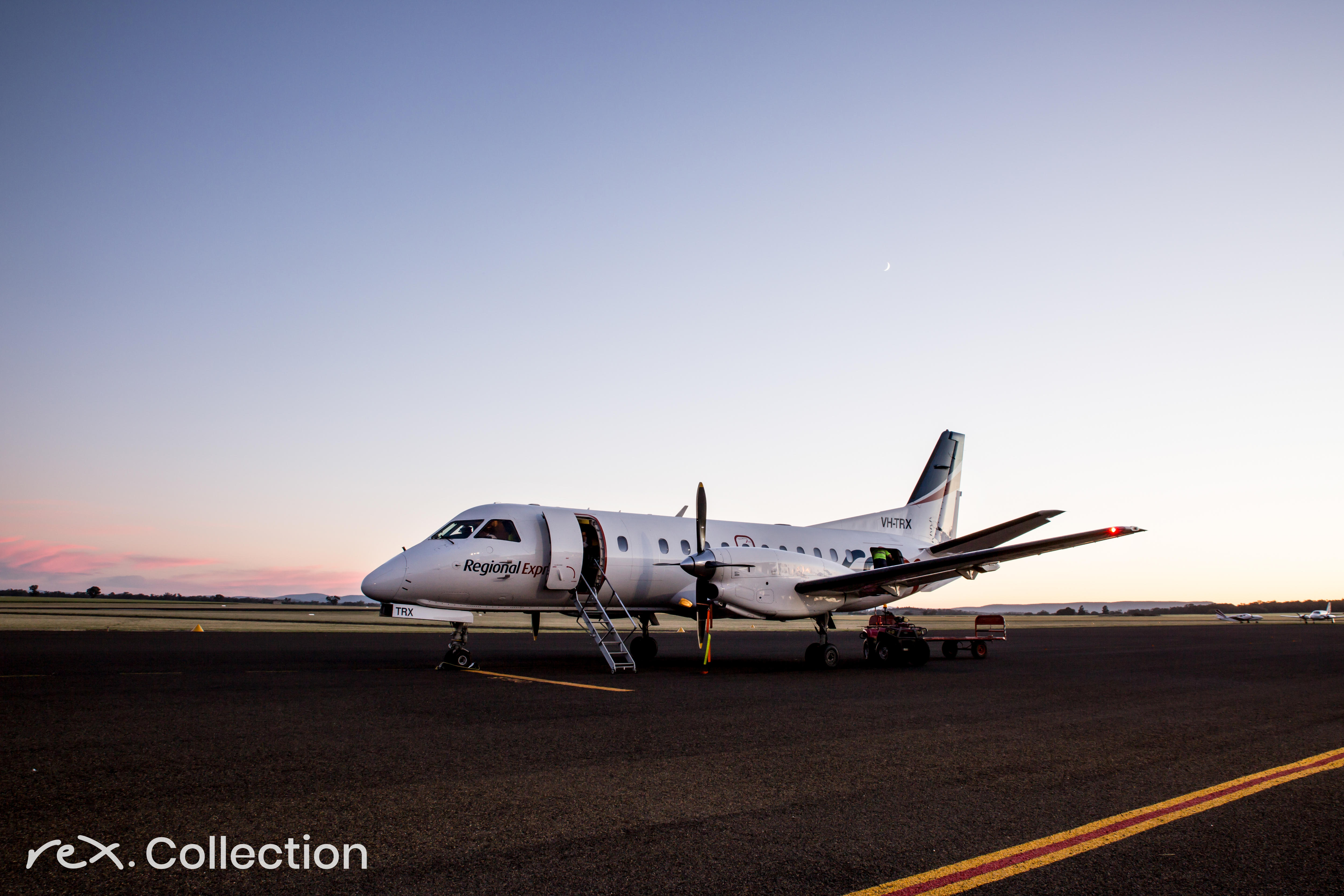 propeller airplane with REX logo sits on tarmac at dusk.