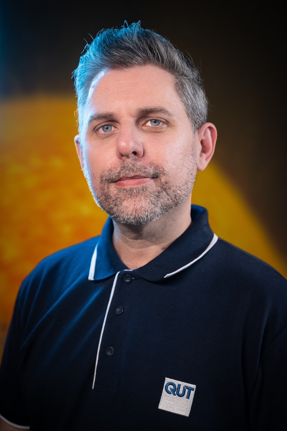 A man in a blue collared shirt standing in front of a yellow and black background.