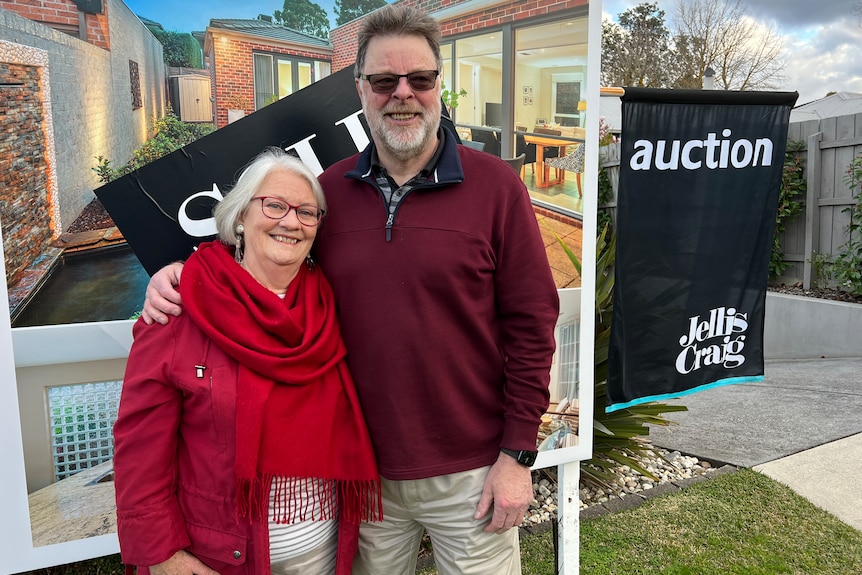 An older man and woman smile in front of a billboard showing a house that had been sold at an auction.