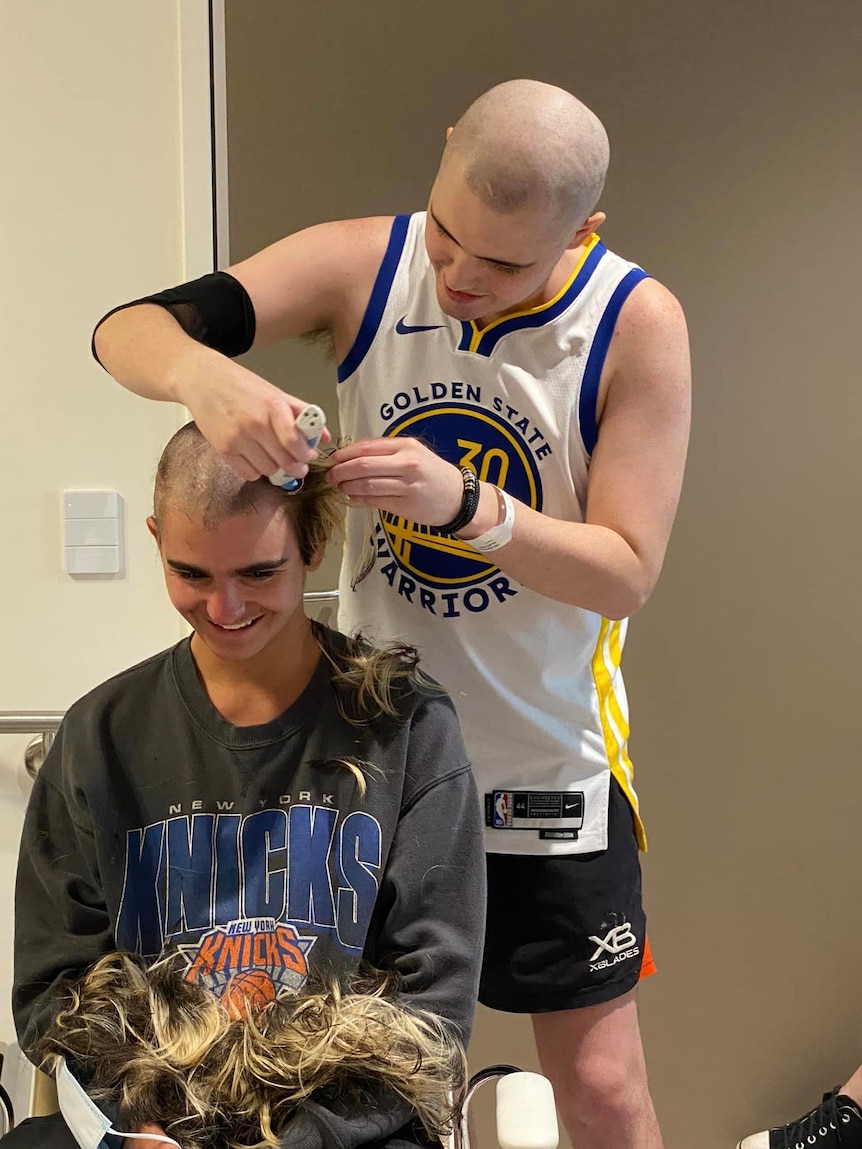 A young man wearing a basketball outfit with a shaved head shaving the head of another young man sitting