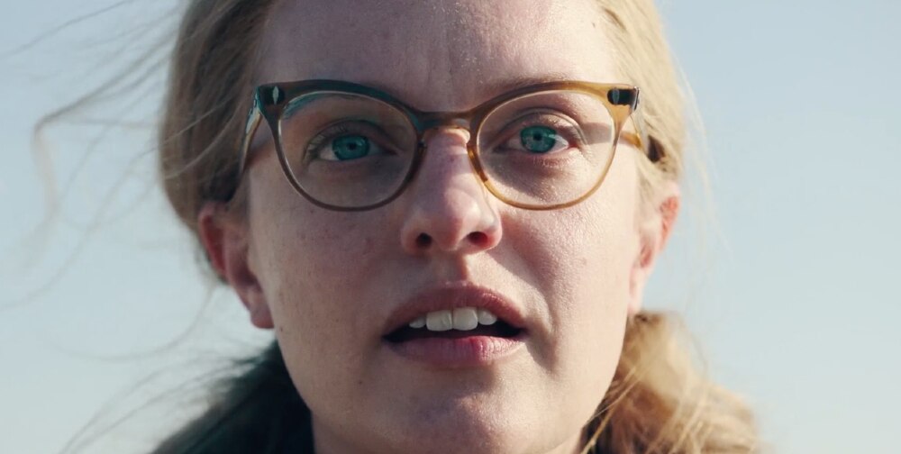 Blue-eyed bespectacled woman's pulled back blonde hair has blank expression on clear sky day, her face half lit by the sun.