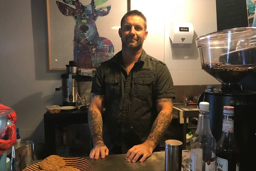 A man standing behind a cafe counter, with a plate of cookies in front of him.