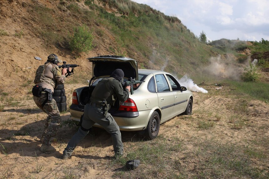 Two combatants in fatigues shoot rifles from behind a car towards a smoke grenade, using the car's raised boot as cover.