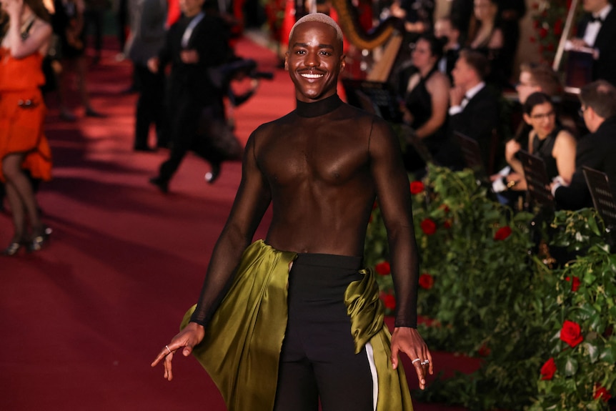 A black man with close cropped bleached hair and a sheer top with an olive toned skirt