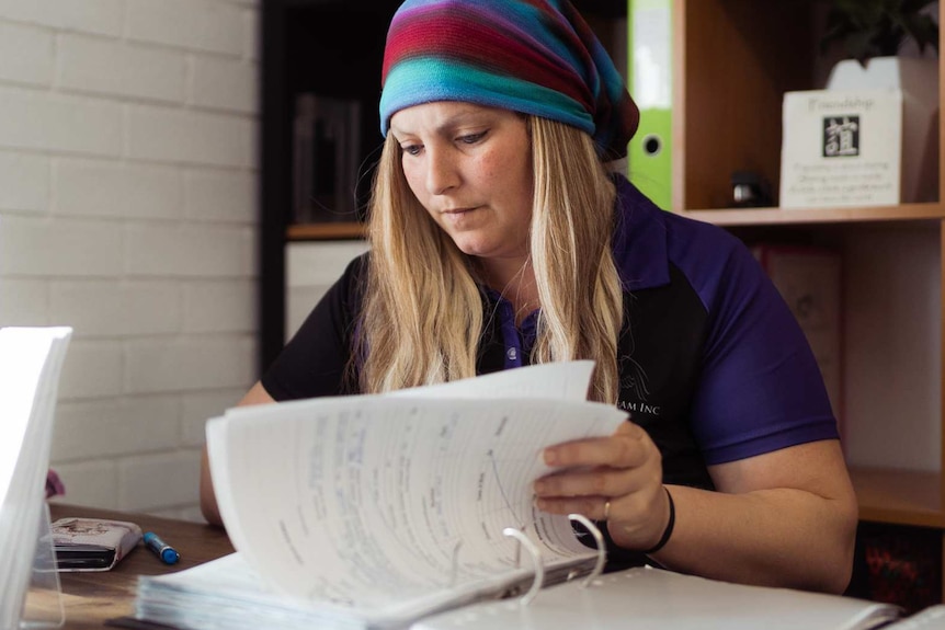 Dee Freitag wearing a black and purple shirt and a striped head scarf sits at a desk looking through a file.