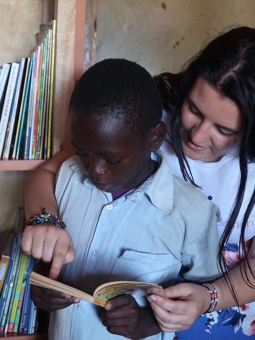 A young Caucasian woman reads a storybook to a young boy of African appearance.