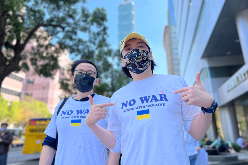 Two young men, shot from slightly below, wear shirts saying NO WAR with the Ukrainian flag on them. One points to his chest 