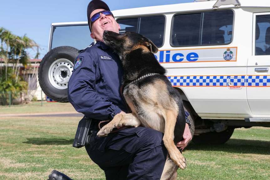 A police officer kneels on the grass, with his police dog jumping up onto him.