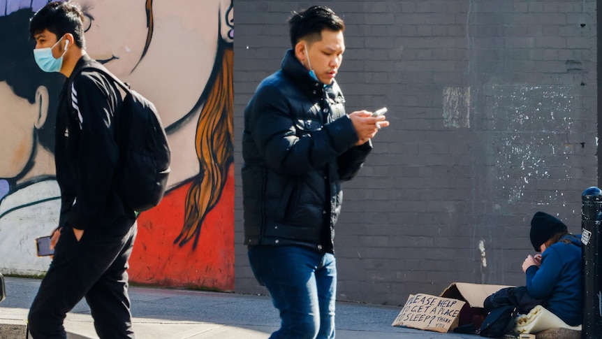 A person sits on the street while two others walk around them.