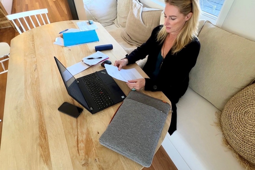 A woman with blonde hair sits at a table with a laptop and documents strewn about. She is reading something on a piece of paper.