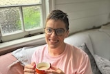 An image of a woman wearing glasses, a pink shirt, sitting on a couch, holding a mug, with a window and wall in the background.