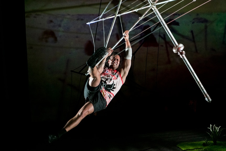 Colour photo of Kyle Shilling hanging from suspended Hills Hoist on the set of Man with the Iron Neck.