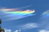 A wispy, rainbow coloured cloud stretches across a bright blue sky, surrounded by sparse white clouds.