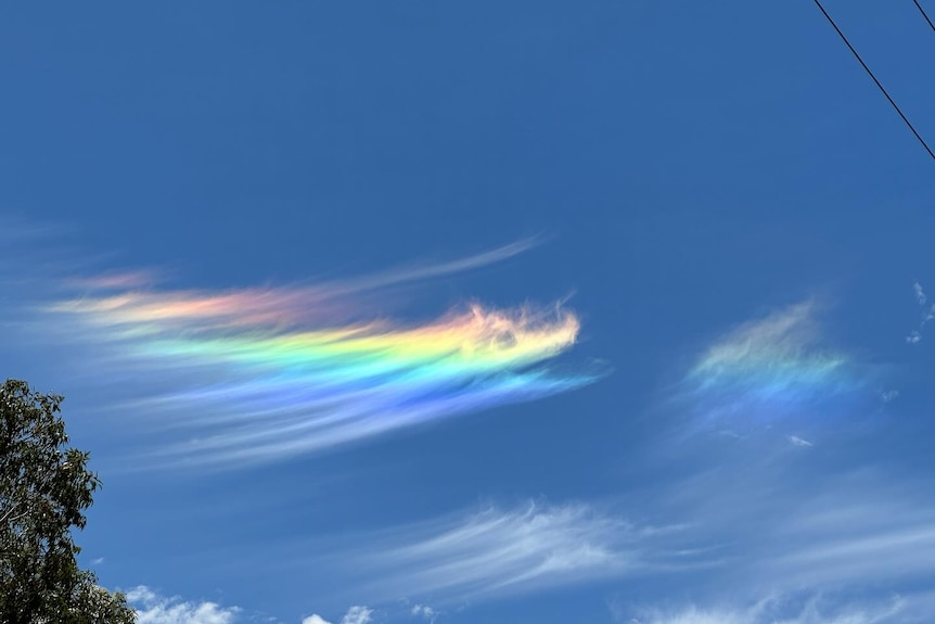 A wispy, rainbow coloured cloud stretches across a bright blue sky, surrounded by sparse white clouds.