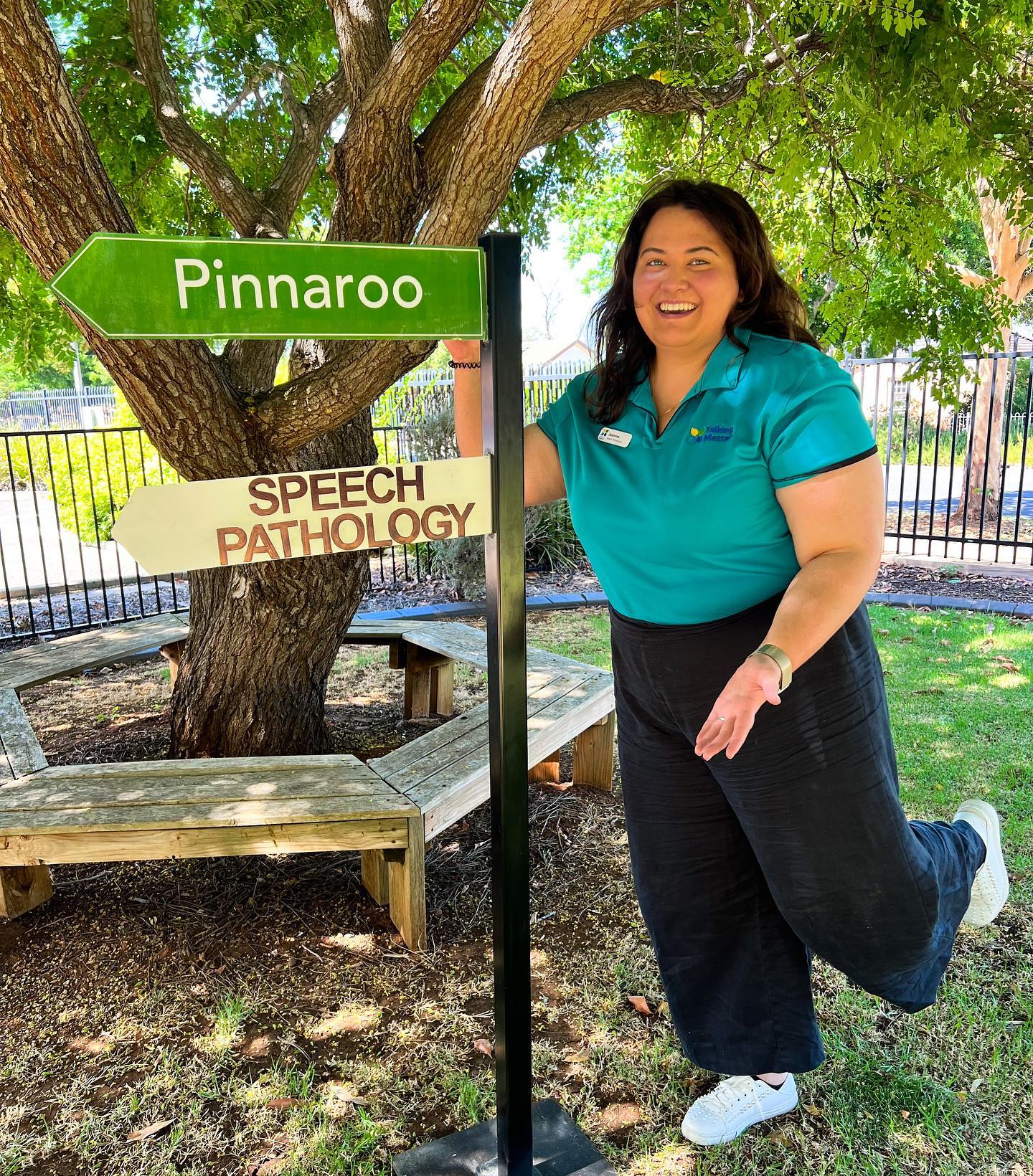 A woman with brown hair, black pants and a teal shirt smiles in front of a sign directing towards pinnaroo