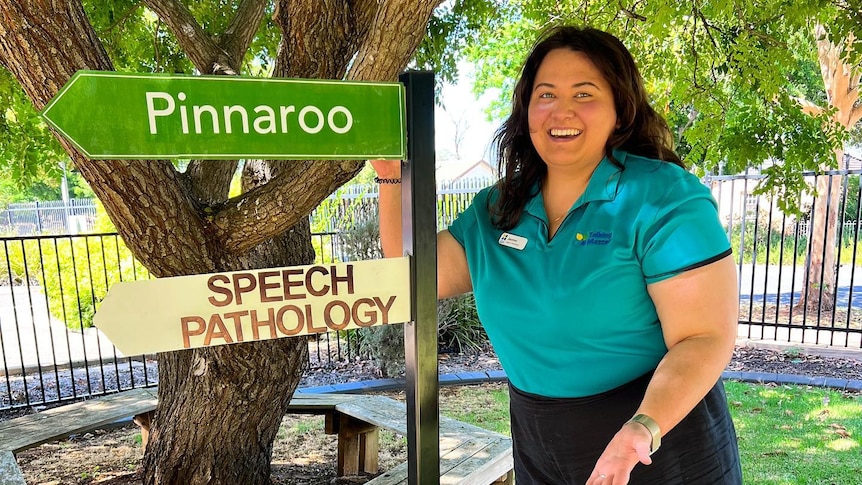 A woman with brown hair, black pants and a teal shirt smiles in front of a sign directing towards pinnaroo