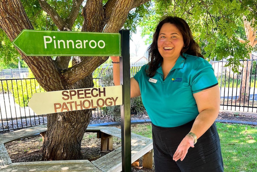 A woman with brown hair, black pants and a teal shirt smiles in front of a sign directing towards pinnaroo