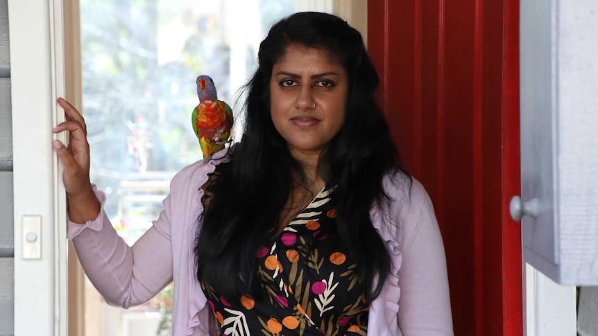 A woman with a rainbow lorikeet on her shoulder looking down the barrel of a camera